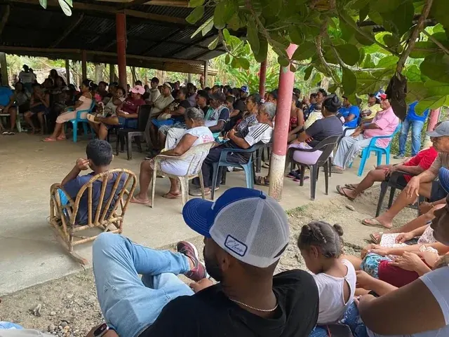 Vecinas y vecinos reunidos bajo un techo abierto junto a árboles.