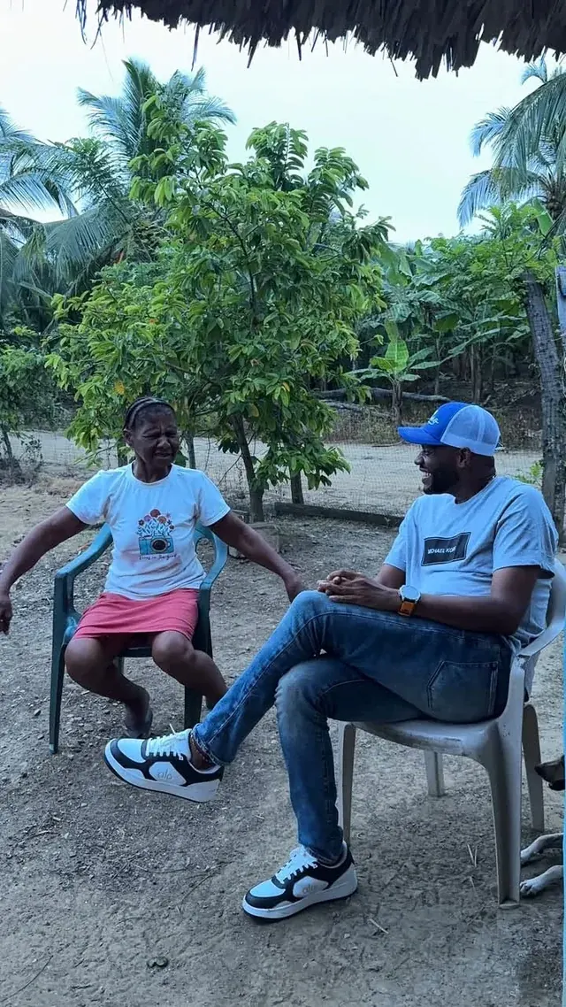 Dos personas conversan sentadas en un patio con árboles y palmeras.
